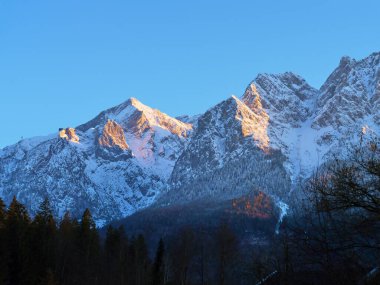 Karla kaplı zirveler, parlak mavi gökyüzünün altındaki ağaçlarla çevrili dağ sırasının üzerinden doğan güneş gibi sabahın ilk ışıklarını yansıtır..