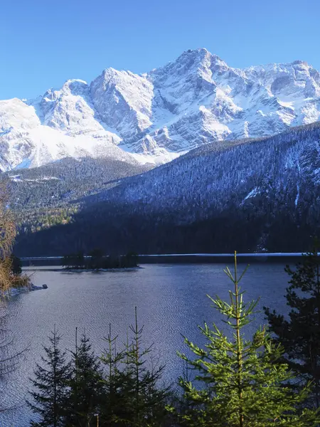 Sakin bir gölün üzerindeki karla kaplı tepeler kışın güzelliğini yansıtıyor. Evergreen ağaçları, aydınlık bir günde sakin atmosferi güçlendiren sahneyi çerçeveler..