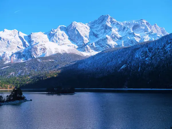 Sakin bir göl üzerindeki görkemli karla kaplı tepeler mavi gökyüzünü ve etrafındaki ormanı yansıtıyor ve gündüzleri huzurlu bir kış ortamı yaratıyor..