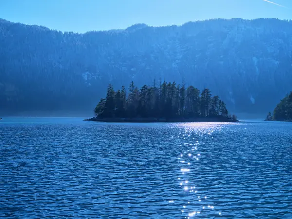 Açık mavi suyla çevrili küçük bir adanın huzurlu görüntüsü güneş ışığını yansıtır. Görkemli dağlar parlak bir gökyüzünün altında arka planda yükseliyor ve sakin bir atmosfer yaratıyor..