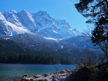 Sakin bir göl üzerinde kar kulesiyle kaplı görkemli dağlar berrak mavi gökyüzünü yansıtıyor. Çam ağaçları kıyı şeridini çerçeveleyerek bu güzel manzaranın huzurlu atmosferini güçlendiriyor..