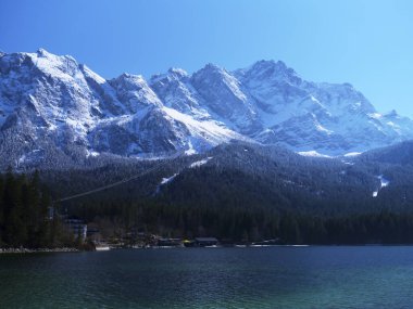 Karla kaplı çarpıcı bir dağ sırası sakin bir göl üzerinde görkemli bir şekilde yükselir. Güneş ışıl ışıl parlıyor, manzarayı aydınlatıyor. Kıyı boyunca uzanan ağaçlar manzarayı güzelleştiriyor..