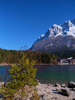 Sakin bir göl, aydınlık ve güneşli bir günde çevredeki dağları ve ormanı yansıtır. Çam ağaçları suların kenarında durur, doğal ortamın huzurlu atmosferini arttırır..
