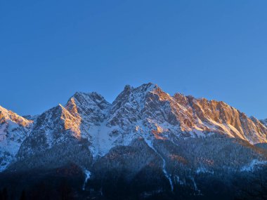 Karla bezenmiş görkemli dağ zirveleri sıcak günbatımı renklerini yansıtır. Gökyüzü açık ve mavidir. Huzurlu bir alp atmosferi yaratır. Doğa güzelliği izleyiciyi cezbeder..