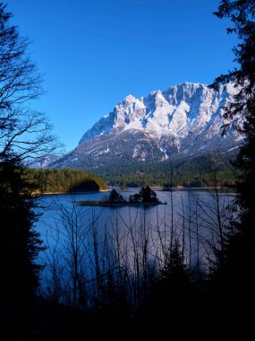 Ağaçlarla çevrili sakin bir göl üzerinde görkemli bir dağ sırası yükseliyor ve berrak mavi bir gökyüzü sergiliyor. Kış manzarası dokunulmamış doğayı yansıtır güzellik ve sakinlik..