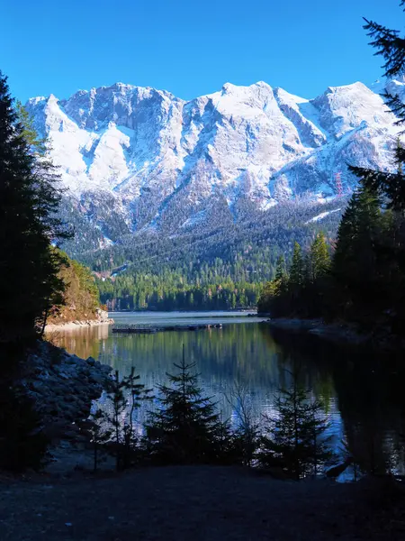 Karla kaplı zirveler, sakin bir gölde yansıtılan manzaraya hükmediyor. Sonsuza dek yeşil ağaçların etrafında, açık mavi bir gökyüzü altında, gün ortasında huzurlu bir ortam sağlar..