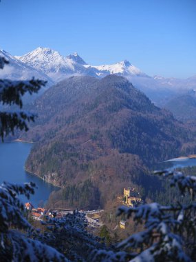 Arka planda görkemli dağlar yükselir, karla kaplı tepeler sergilenir. Sakin bir göl, ağaçların arasına kurulmuş büyüleyici bir kale dururken berrak mavi gökyüzünü yansıtır..