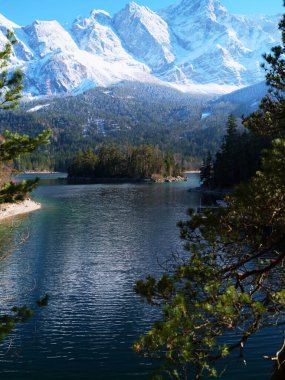 Karla kaplı dağlar, güzelliklerini yansıtan sakin bir gölün üzerinde görkemli bir şekilde yükselir. Bereketli, yemyeşil ağaçlar suyu çerçeveletip açık havada huzurlu bir atmosfer yaratıyor..