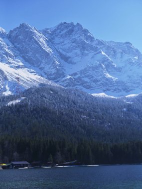 Görkemli kar örtülü dağlar, sonsuz yeşil ağaçlarla çevrili sakin bir göle yansıyan dik dururlar. Parlak mavi gökyüzü huzurlu kış atmosferini güçlendirir..