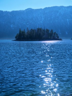 Sakin bir gölün yüzeyinde gün ışığı dans eder. Berrak bir günde, arka planda gür ağaçlar ve görkemli dağlarla çevrili bir adanın altını çizer..