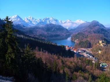Karla kaplı zirveler, mavi gökyüzünü yansıtan sakin bir göl üzerinde görkemli bir şekilde yükselirken, yemyeşil ormanlarla çevrili dağ eteklerinde tuhaf bir köy dinleniyor..