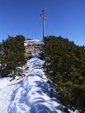 Dağların tepesinde, yemyeşil ağaçlarla çevrili ahşap bir haça giden kayalık yol karla kaplıdır. Açık mavi bir gökyüzü huzurlu atmosferi güçlendirir.