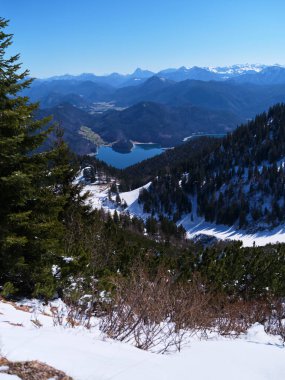 Bir dağ zirvesinden nefes kesici bir manzara. Karla kaplı tepeler arasında uzanan donmuş bir göl. Ve açık mavi bir kış günü açık mavi gökyüzünün altında yemyeşil ağaçlar..