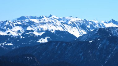 Karla kaplı uzak tepeler parlak mavi gökyüzüne karşı görkemli bir şekilde yükselir. Engebeli dağ arazisinde çeşitli yamaçlar sergileniyor. Doğal güzellik ve kış dinginliği vurgulanıyor..