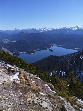 Kayalık bir dağın tepesinde, göz kamaştırıcı bir manzara, parlak güneş ışığı altında karla kaplı dağların çerçevelediği, gökyüzünü yansıtan sakin bir gölü gözler önüne seriyor. Doğanın güzelliği gözler önünde..