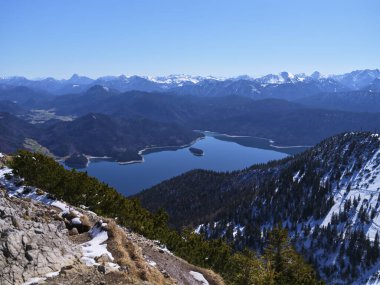 Karla kaplı tepeler ve Bavyera Alpleri 'ndeki açık mavi gökyüzünün altında yemyeşil bitki örtüsüyle çevrili pırıl pırıl bir göl nefes kesici manzaralar sergiliyor..