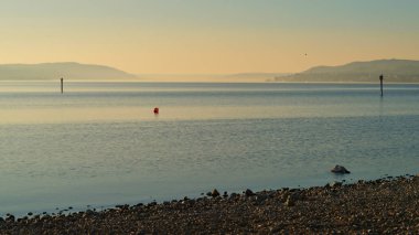 Sabahın erken saatlerinde sakin suların üzerinde altın rengi belirir, çakıl taşları ve uzak tepelerle benekli bir kıyı şeridini aydınlatır. Bir şamandıra suyu işaretliyor, dingin atmosfere ekliyor..