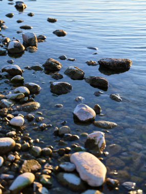 Yumuşak taşlar ve çakıl taşları, gün batımının yumuşak ışığını yansıtan huzurlu bir gölün kıyı şeridini oluşturur. Sakin su yavaşça kayalara çarpar ve sakin bir atmosfer yaratır..