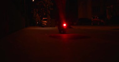 Bir insan gece loş bir şehir caddesinde scooter kullanıyor. Parlayan arka ışık, etrafındaki karanlıkla çelişiyor. İlerideki yolu aydınlatıyor..