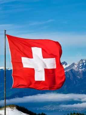 Rüzgarda dalgalanan beyaz haçlı kırmızı bir bayrak, İsviçre dağlarının çarpıcı zemininde ve açık mavi gökyüzünde. Atmosfer canlı ve huzurlu..