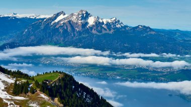 Karla kaplı dağlar yemyeşil bir vadi ve göl üzerinde belirgin bir şekilde yükselir. Parlak bir bahar gününde, bulutlarla çevrili bir köy manzaranın içinde sıkışıp kalmış..