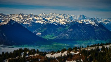 Yüksek bir bakış açısından, kar kaplı dağların nefes kesici manzarası ufuk boyunca uzanır. Altta yemyeşil vadiler uzanır, doğada çarpıcı bir kontrastlık yaratır..
