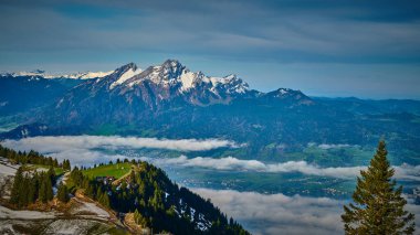 Karla kaplı zirveler, altında ince bulutlar yüzerken yemyeşil vadilerin üzerinde görkemli bir şekilde yükselir. Parlak gün ışığı, İsviçre manzarasının doğal güzelliğini güçlendiriyor..