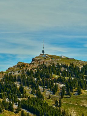 Bir iletişim kulesi, her yer yeşil ağaçlar ve yuvarlanan tepelerle çevrili bir dağın zirvesinde dimdik durur. Sahne, parlak mavi gökyüzünün altında canlı bir manzara sergiliyor..