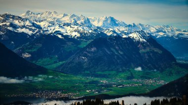 Karla kaplı zirveler, yemyeşil vadiler ve yumuşak yamaçlarla çevrili dramatik bir şekilde yükselir. Sakin atmosfer sabahın erken saatlerindeki ışığı yansıtıyor ve çarpıcı bir doğal manzara yaratıyor..