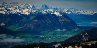 Karla kaplı zirveler, bulutlu bir gökyüzünün altındaki küçük köylerle benekli yemyeşil bir vadide görkemli bir şekilde yükseliyor. Atmosfer sakin ve nefes kesici..