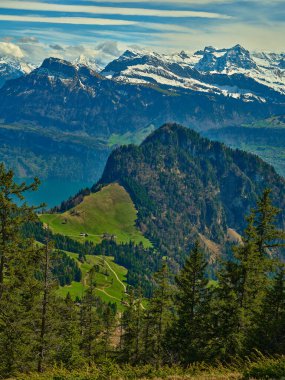 Görkemli dağlar yemyeşil tepelerin üzerinde, dingin bir göl tepeleri yansıtırken. Açık mavi gökyüzü bu dağlık manzaranın güzelliğini gün ışığında artırır..