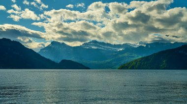 Sakin bir gölü çevreleyen görkemli dağlar yukarıdaki bulutları yansıtıyor. Bereketli yeşillik kıyıları kaplıyor. Gün ortasında sakin bir atmosfer yaratıyor..