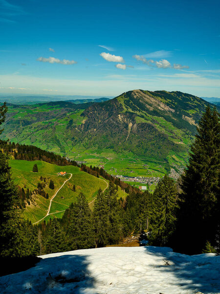 Spectacular landscape featuring rolling green hills and mountain ranges under a clear blue sky, with snow still visible in shaded areas, highlighting the beauty of nature in springtime.