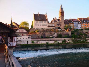Gece ışığı nehir kenarındaki antika kasabayı yıkıyor, tarihi mimariyi ve taş kuleyi aydınlatıyor. Ahşap bir köprü, akan su ve çevreyi manzaralı bir şekilde gösterir..