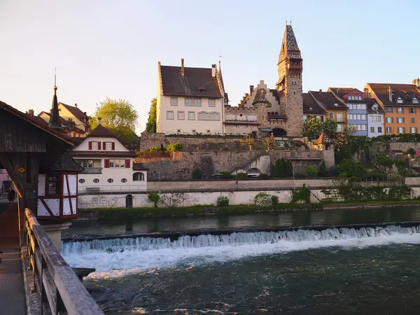 Gece ışığı nehir kenarındaki antika kasabayı yıkıyor, tarihi mimariyi ve taş kuleyi aydınlatıyor. Ahşap bir köprü, akan su ve çevreyi manzaralı bir şekilde gösterir..