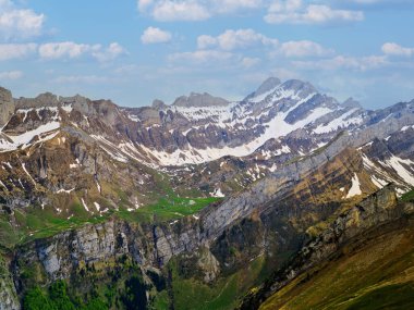 Karla kaplı zirveler yeşil vadilerin üzerinde görkemli bir şekilde yükselir, açık bir günde dağların güzelliğini gözler önüne serer. Kabarık bulutlar engebeli arazinin üzerinde süzülür ve manzarayı zenginleştirir..