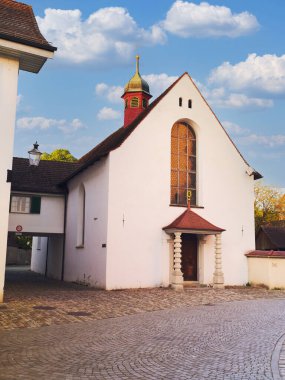 Mavi gökyüzünün altında sakin bir köy atmosferiyle çevrili, kendine özgü bir çan kulesi ve geniş kemerli penceresi olan, tuhaf beyaz bir kilise kaldırım taşı sokağının yanında duruyor..