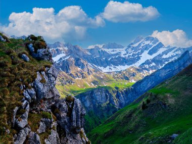 Karla kaplı dağlar, yemyeşil vadilerin üzerinde, nefes kesici bir manzara yaratıyor. Açık mavi gökyüzü pofuduk bulutlarla kaplı, manzarayı canlı ve huzurlu kılıyor..