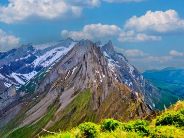 Karla kaplı zirveler, yemyeşil çayırlarla çevrili canlı mavi gökyüzüne çarpıcı bir şekilde yükselir. Manzara, yazın doğanın katıksız güzelliğini gözler önüne seriyor..