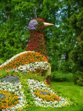 Çiçeklerden yapılmış büyük, renkli bir kuş heykeli yeşil bir bahçenin ortasında duruyor ve bir dizi çiçeğin uzun ağaçların önünde sergileniyor. Doğa sanatları tamamen sergileniyor..