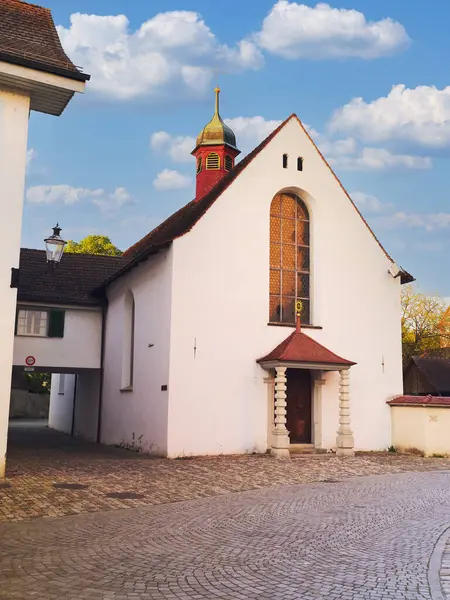 Mavi gökyüzünün altında sakin bir köy atmosferiyle çevrili, kendine özgü bir çan kulesi ve geniş kemerli penceresi olan, tuhaf beyaz bir kilise kaldırım taşı sokağının yanında duruyor..
