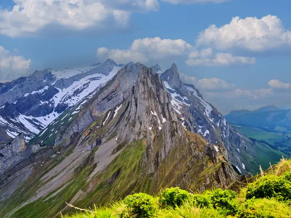 Karla kaplı zirveler, yemyeşil çayırlarla çevrili canlı mavi gökyüzüne çarpıcı bir şekilde yükselir. Manzara, yazın doğanın katıksız güzelliğini gözler önüne seriyor..