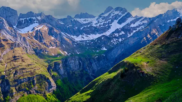 Güçlü dağlar parlak bir gökyüzüne karşı görkemli bir şekilde yükselir, karla kaplı zirveler ve zengin yeşil vadiler sergilerler. Manzara, güzelliği huzurlu bir anda yakalar..