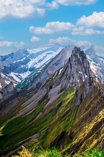 Karla kaplı yamaçlar ve kabarık beyaz bulutların altında canlı yeşil tepelerle çevrili gökyüzüne doğru yükselen yüksek dağ zirveleri..