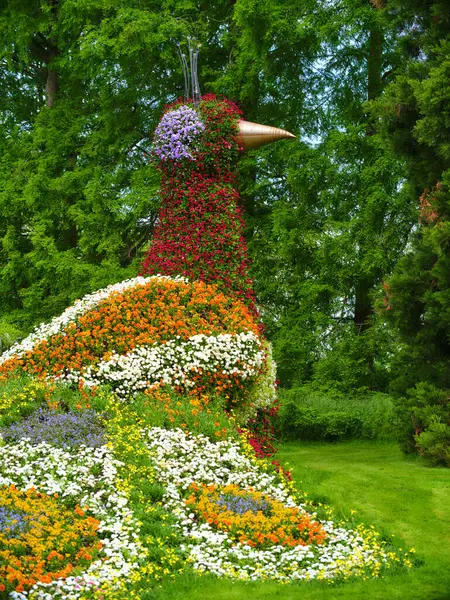 Çiçeklerden yapılmış büyük, renkli bir kuş heykeli yeşil bir bahçenin ortasında duruyor ve bir dizi çiçeğin uzun ağaçların önünde sergileniyor. Doğa sanatları tamamen sergileniyor..