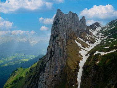Çarpıcı bir dağ zirvesi, yemyeşil tepeler ve kar kalıntıları üzerinde, berrak mavi gökyüzü ve yumuşak bulutlardan oluşan bir zemin üzerinde aydınlanan, güzellikleri gözler önüne seren, güneş ışığıyla aydınlanan..