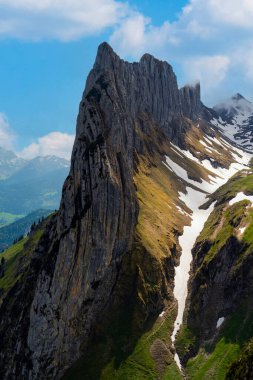 Bereketli vadilerin ve kar yığınlarının üzerinde çarpıcı bir dağ zirvesi, güneşli bir yaz gününde açık mavi gökyüzünün altında doğanın güzelliğini gözler önüne seriyor..