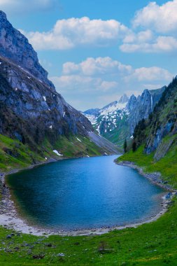 Dik dağ yamaçlarının arasına kurulmuş sakin bir göl canlı mavi gökyüzünü yansıtır. Uzakta karla kaplı tepeler belirirken yemyeşil tepeler suları kucaklıyor..