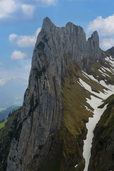 Yeşil yamaçlar ve kar parçalarıyla çevrili açık mavi gökyüzüne doğru yükselen yüksek kaya oluşumu. Doğal güzellik, dağlık arazinin özünü yakalar..