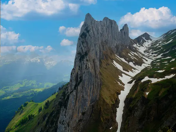 Çarpıcı bir dağ zirvesi, yemyeşil tepeler ve kar kalıntıları üzerinde, berrak mavi gökyüzü ve yumuşak bulutlardan oluşan bir zemin üzerinde aydınlanan, güzellikleri gözler önüne seren, güneş ışığıyla aydınlanan..